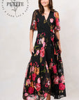 A woman modeling a black maxi dress with a vibrant pink, red, and orange floral print. The dress features sheer flutter sleeves, a V-neckline, and a matching self-tie belt at the waist. Hope & Ivy
