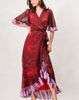 A woman modeling a long wrap dress with a deep red base and a floral print in red, purple, and green. The dress features sheer flutter sleeves with purple ruffled trim, a matching waist tie, and a ruffled hemline. Hope & Ivy