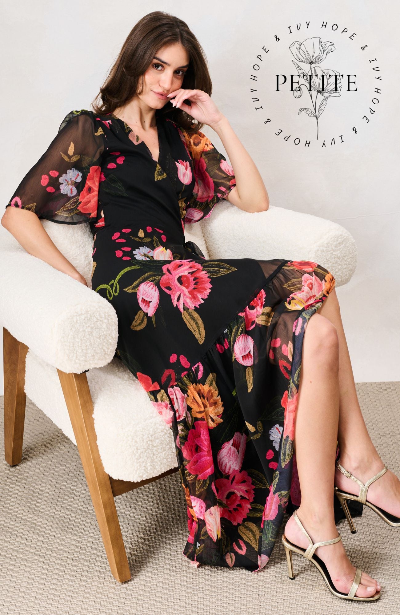 A woman models a black midi dress with a vibrant pink, red, and orange floral print while sitting in a white boucle armchair. The brand logo 'Petite' is visible in the top right corner.