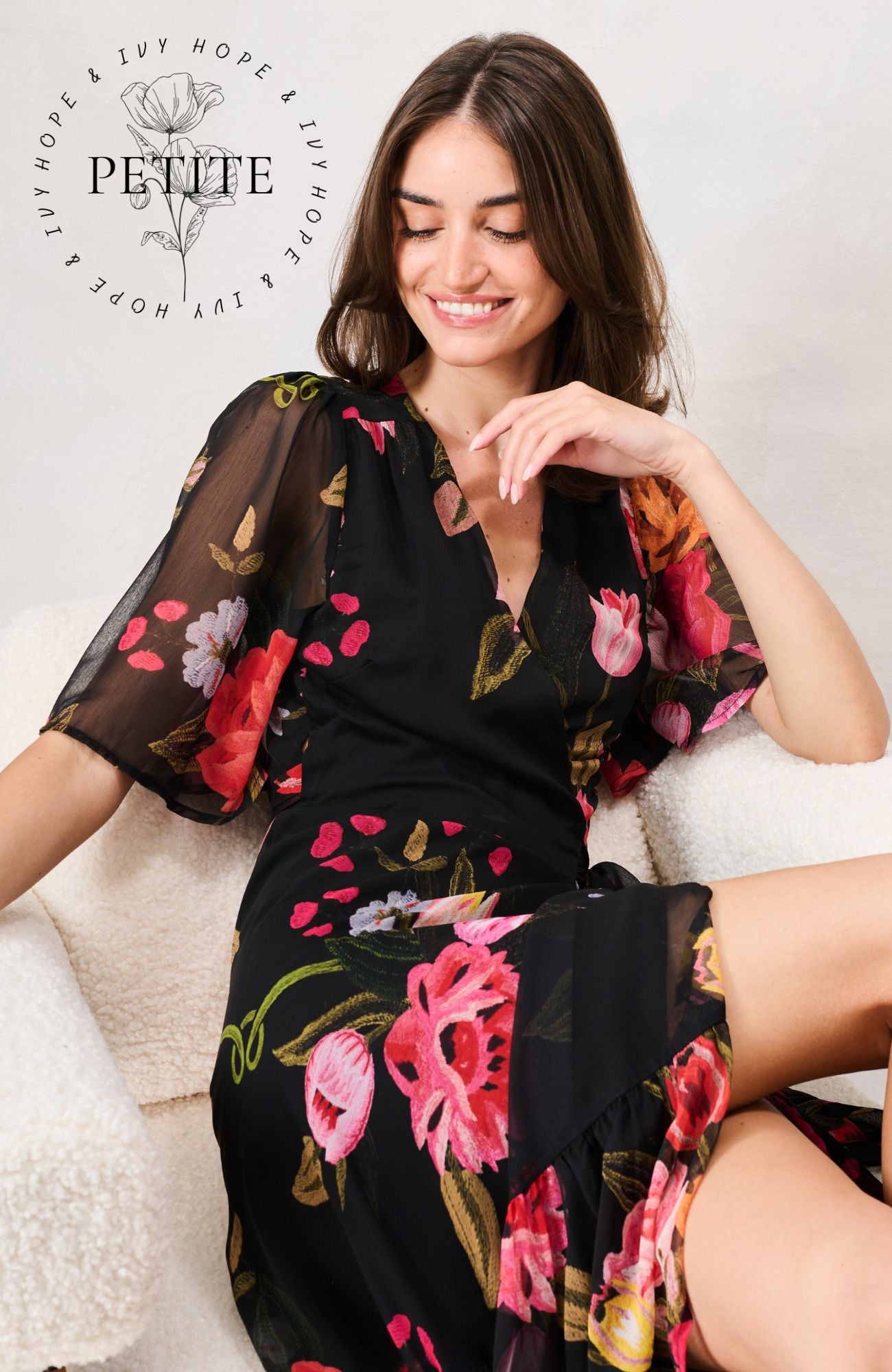 A woman wearing a black floral midi dress with sheer flutter sleeves sits on a white textured sofa. The dress features a vibrant pink, red, and green floral pattern. Hope & Ivy 