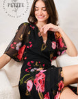 A woman wearing a black floral midi dress with sheer flutter sleeves sits on a white textured sofa. The dress features a vibrant pink, red, and green floral pattern. Hope & Ivy 