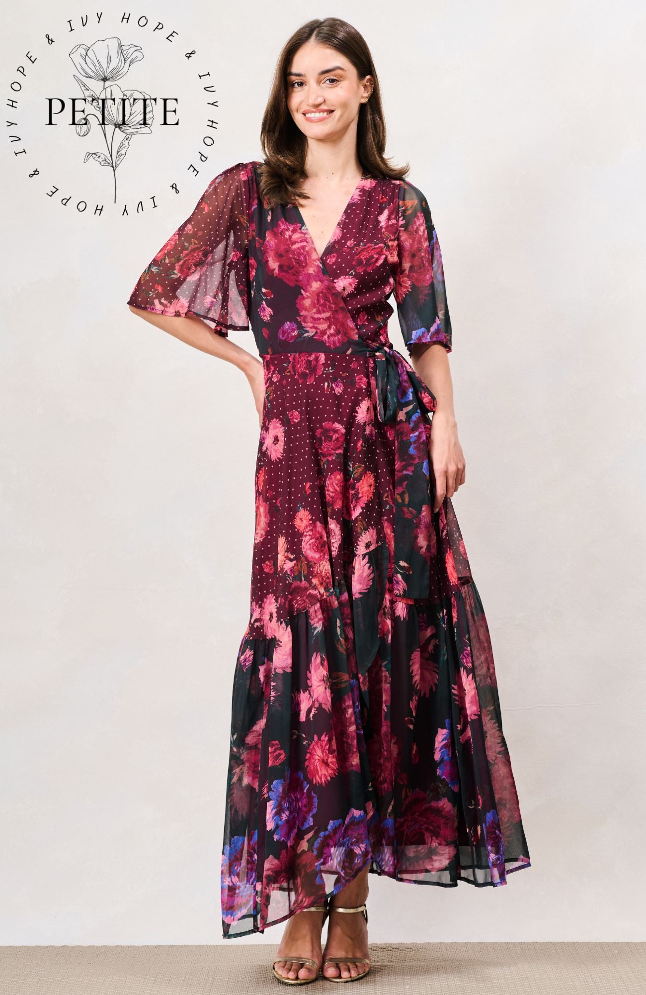 A woman modeling a long floral maxi dress with sheer flutter sleeves and a V-neckline, shown against a plain light background. The dress has a dark base with prominent magenta, pink, and purple floral patterns. Hope & Ivy 