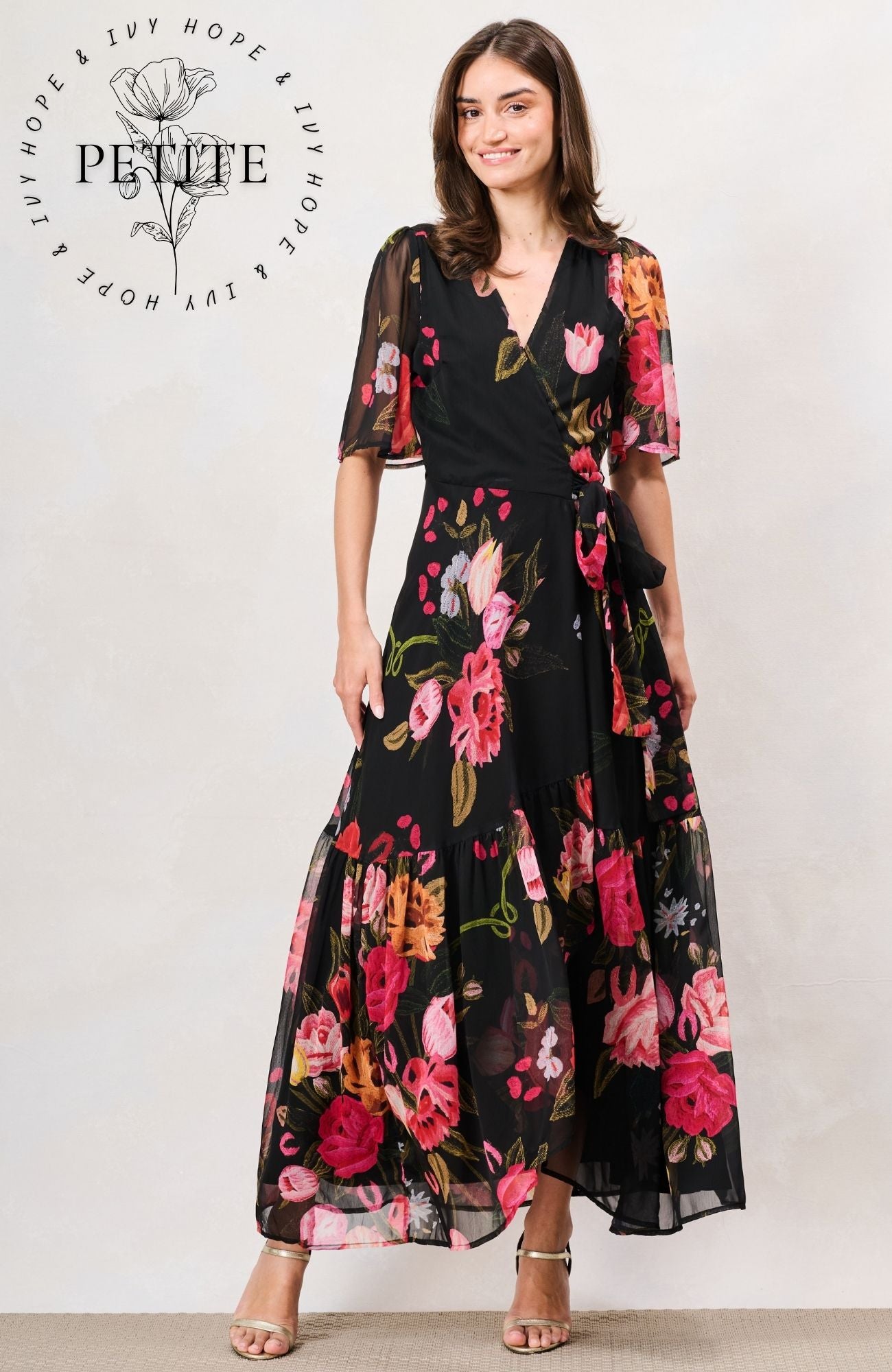 A woman modeling a black maxi dress with a vibrant pink, red, and orange floral print. The dress features sheer flutter sleeves, a V-neckline, and a matching self-tie belt at the waist. Hope & Ivy 