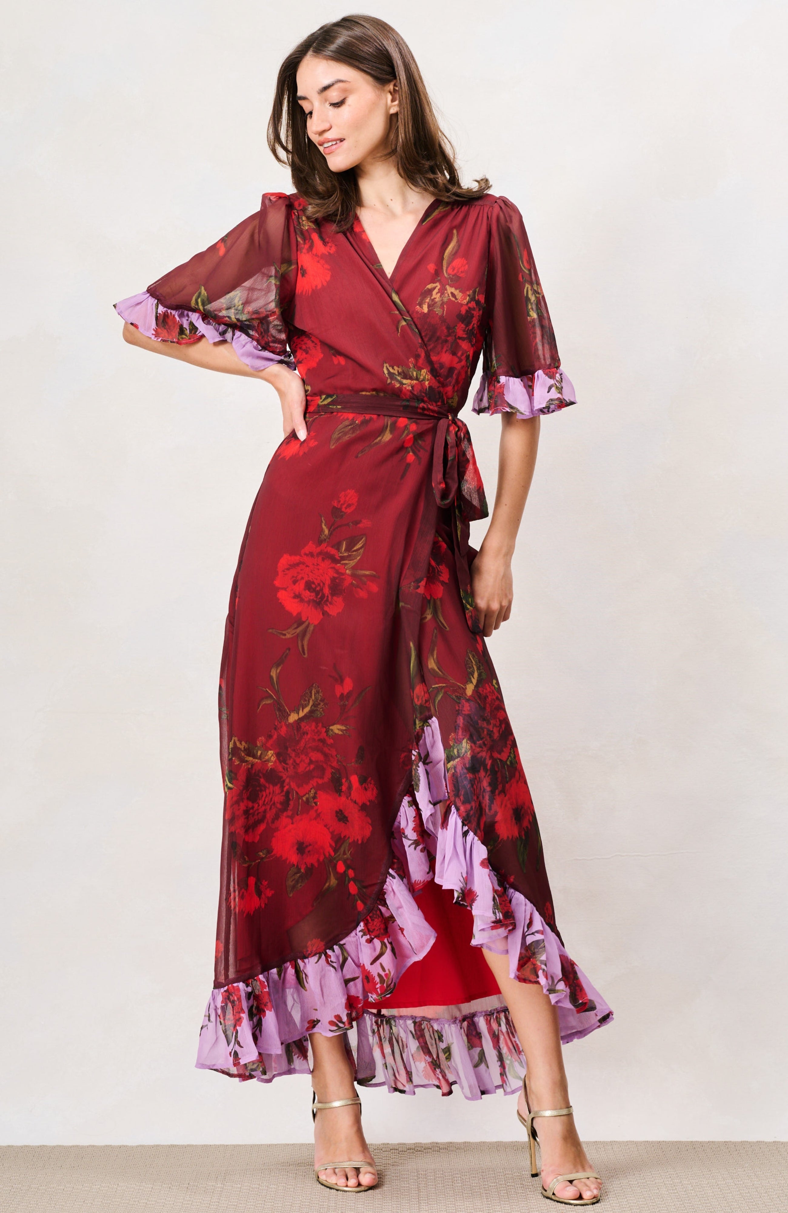 A woman modeling a long wrap dress with a deep red base and a floral print in red, purple, and green. The dress features sheer flutter sleeves with purple ruffled trim, a matching waist tie, and a ruffled hemline. Hope & Ivy 