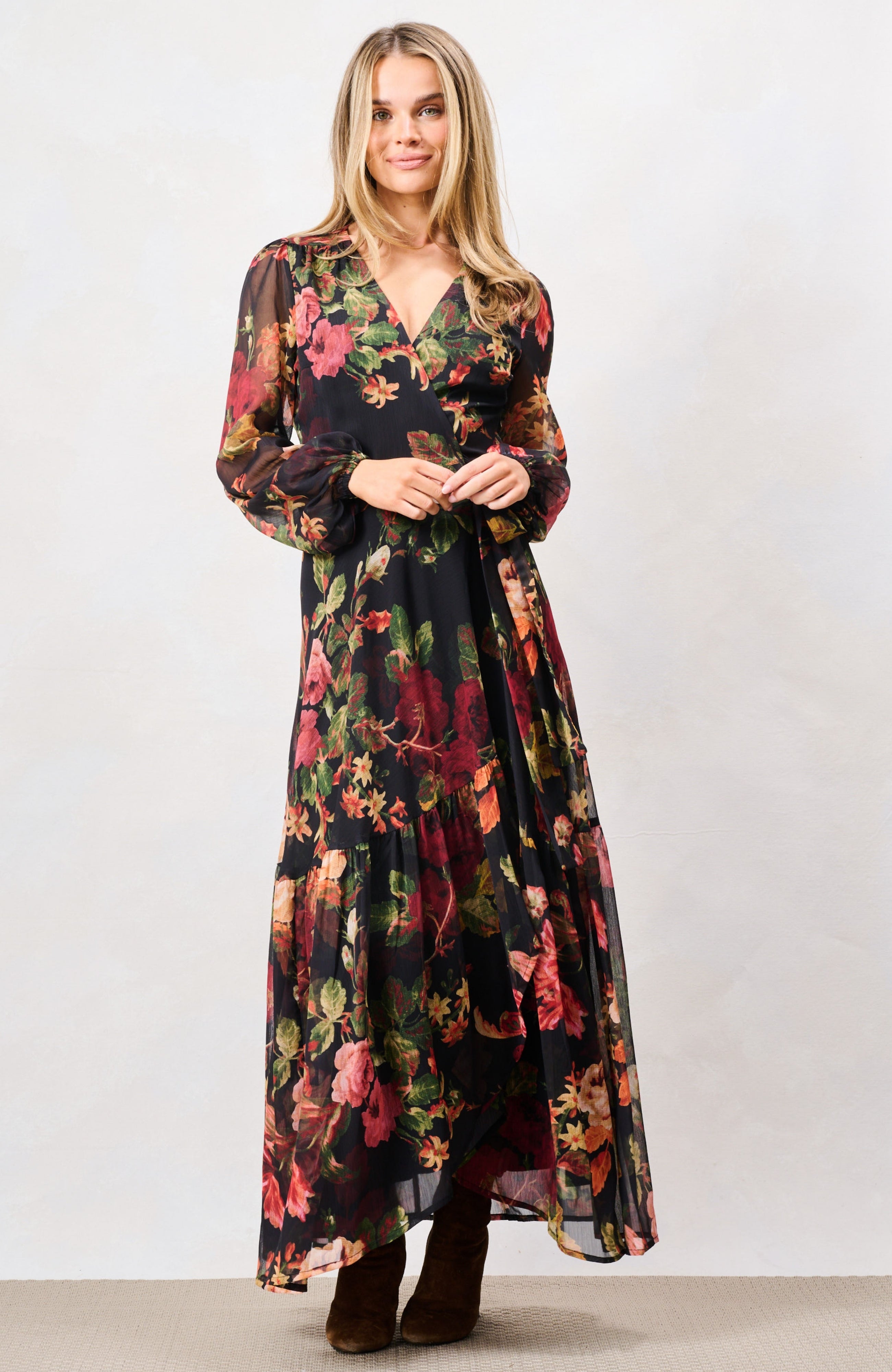 A woman modeling a long-sleeve maxi dress with a dark floral print, featuring a V-neckline and a tie at the waist, paired with dark brown boots against a plain studio background. Hope & Ivy