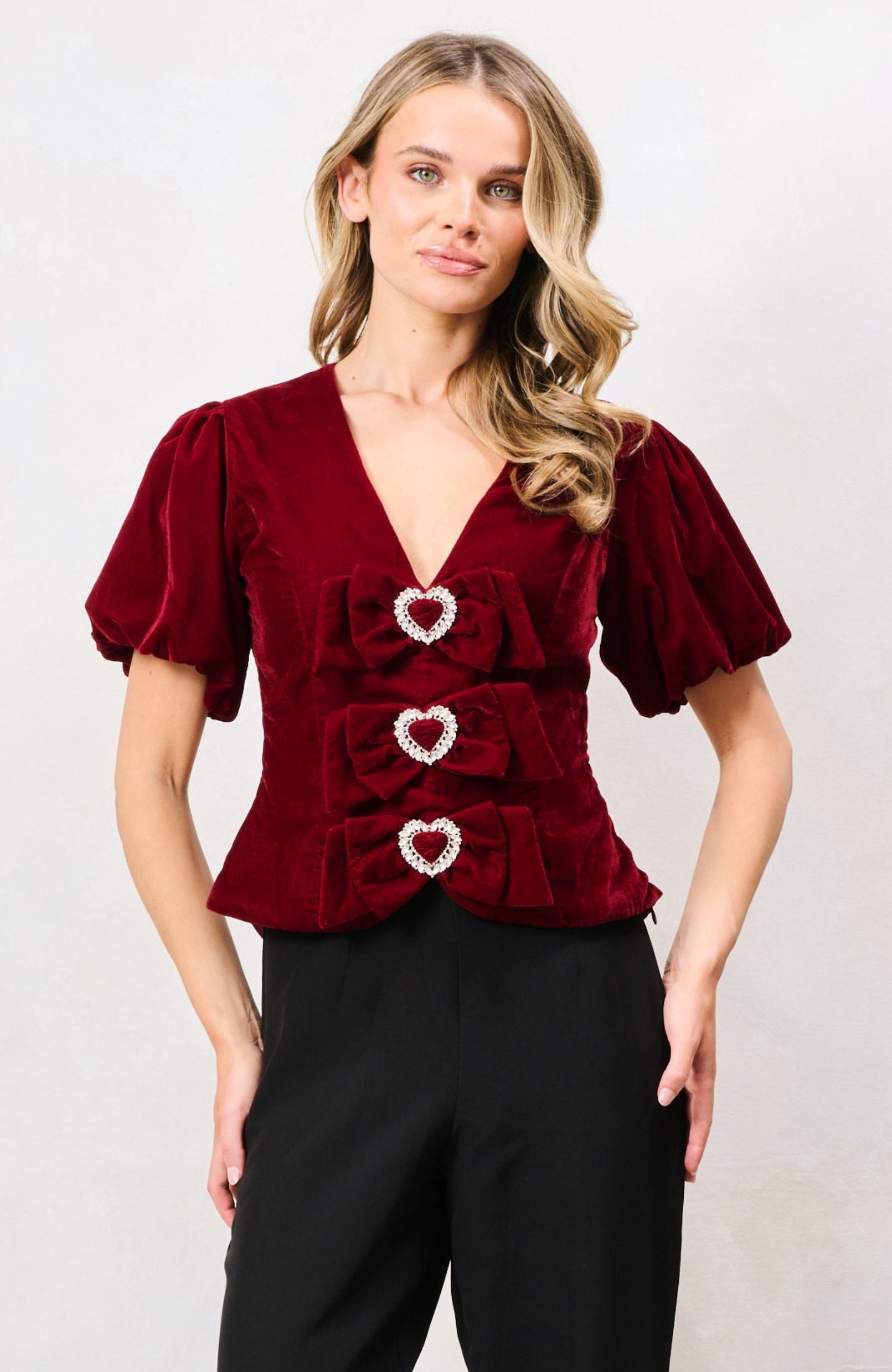 A woman modeling a deep burgundy velvet short-sleeve top with heart-shaped decorative details on the front, paired with black wide-leg trousers, against a plain light gray studio background.