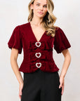 A woman modeling a deep burgundy velvet short-sleeve top with heart-shaped decorative details on the front, paired with black wide-leg trousers, against a plain light gray studio background.